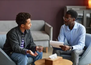 A man and a boy are seated on a couch, discussing topics related to adolescent therapy.