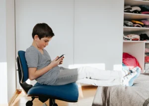A boy sitting on a chair, focused on his cell phone, reflecting concerns about screen time and mental health in youth.